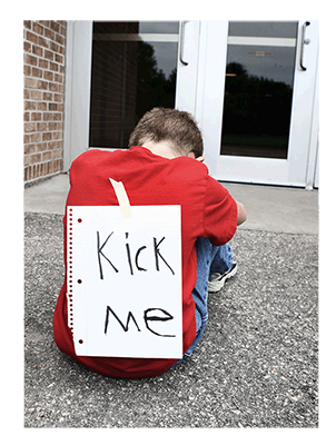 A child outside a school with a "kicj me" sign on his back